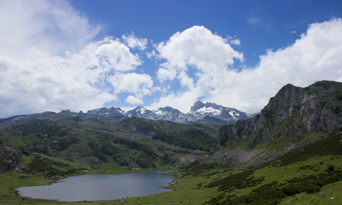 Mountain valley landscape with a calm lake in foreground, green slopes, and snow-capped rocky peaks under a partly cloudy sky