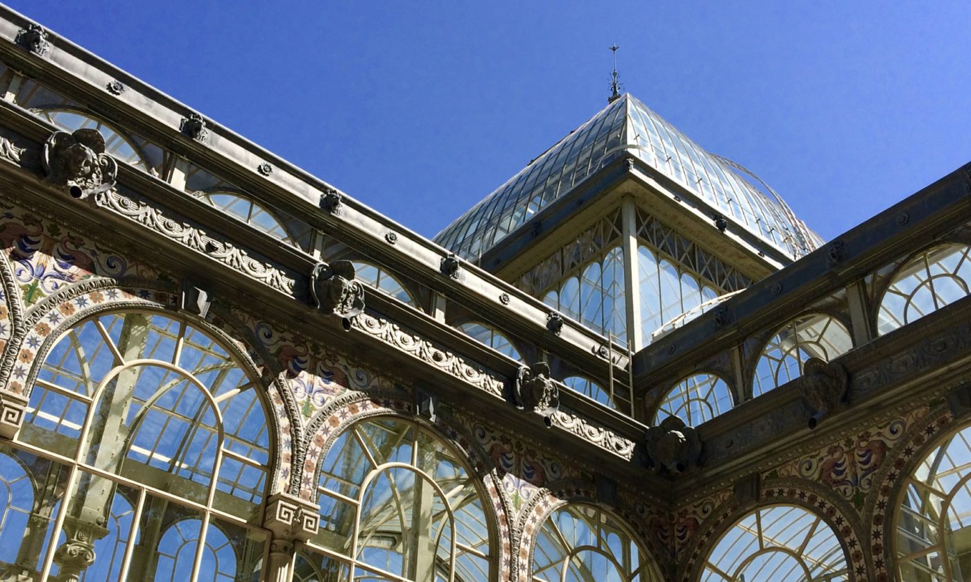 Close-up upward view of an ornate glass and iron pavilion with decorative tilework and a domed glass roof against a clear blue sky