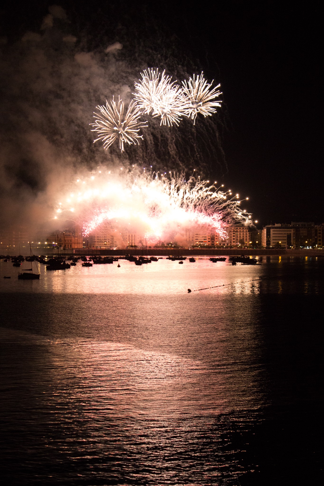 White and pink fireworks bursting over a city skyline at night, reflected on water with boats in foreground