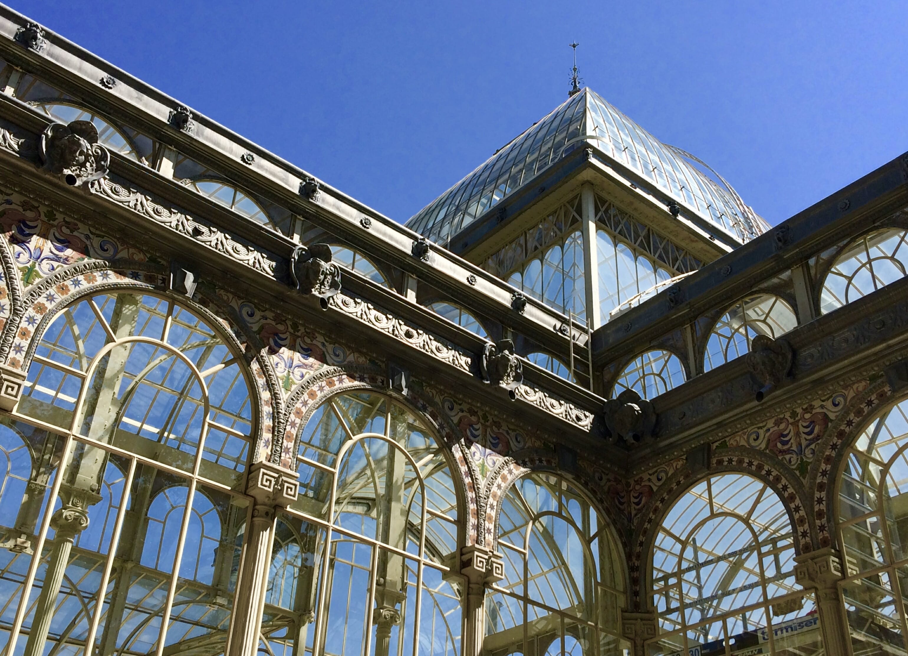 Ornate glass and iron pavilion with domed roof and decorative ironwork, seen against a bright blue sky; elegant, historic architecture