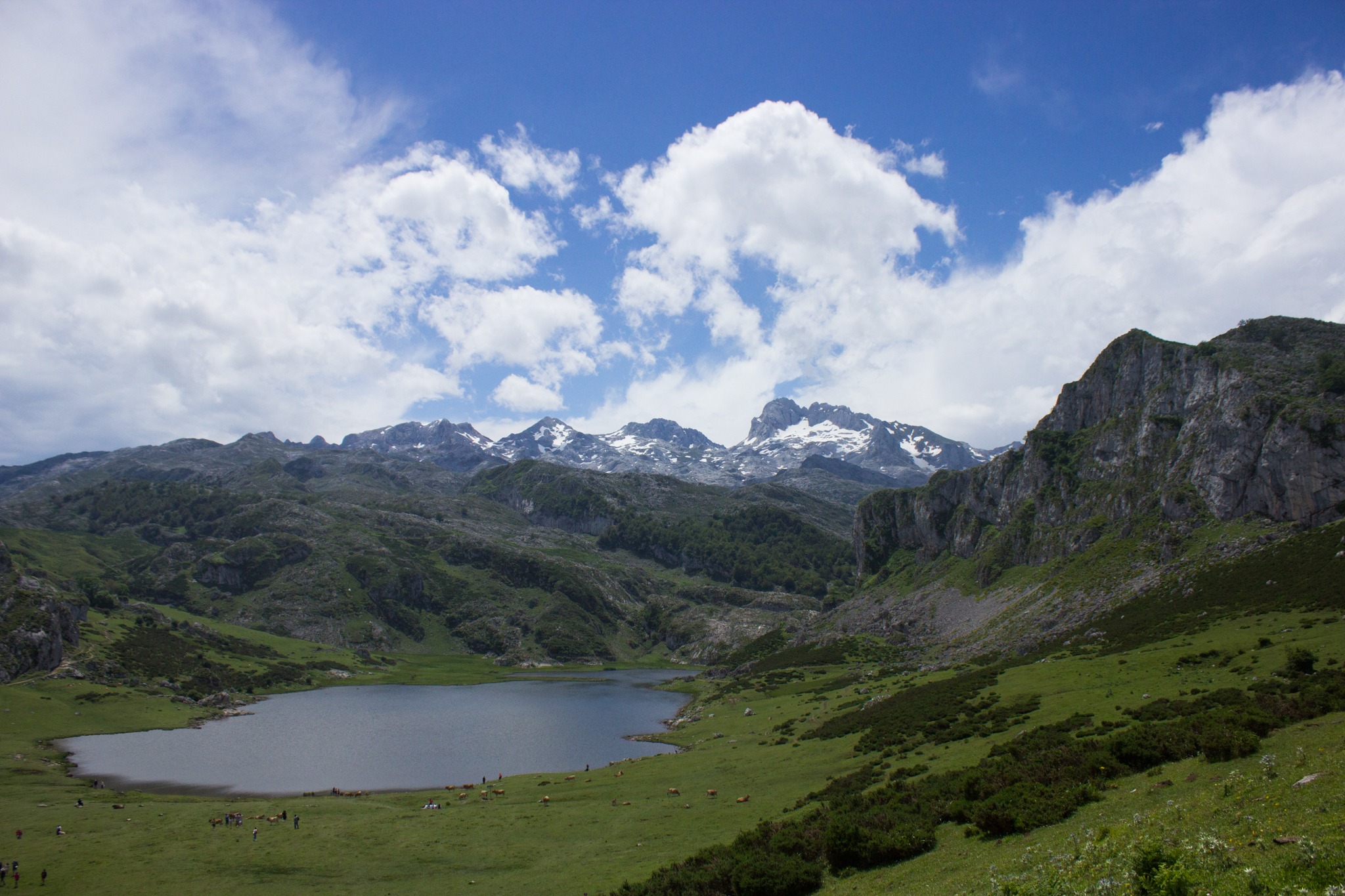 Green valley with a calm lake surrounded by rugged, snow-capped mountains under a partly cloudy sky.