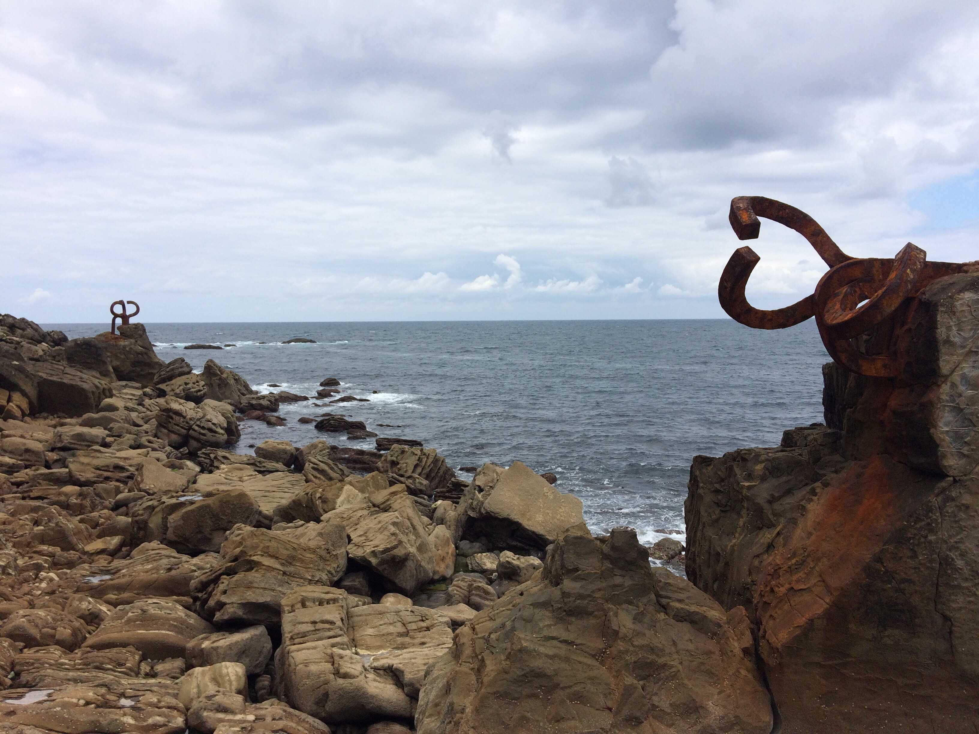 Rocky coastline with dramatic rusted metal sculpture pieces overlooking the sea under a cloudy sky