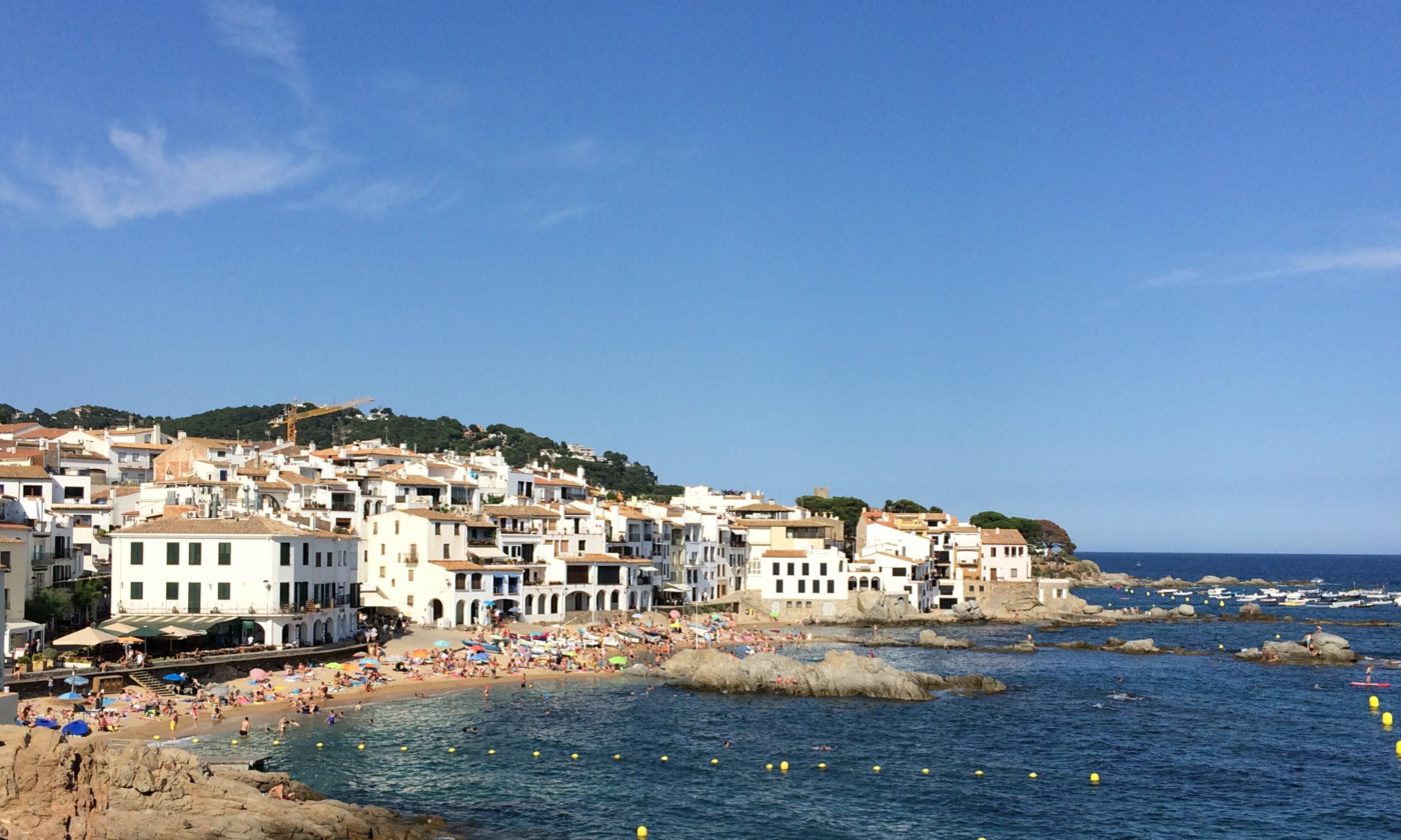 Coastal town with white buildings clustered on a rocky shoreline, a small beach filled with people, and clear blue sea under a bright sky